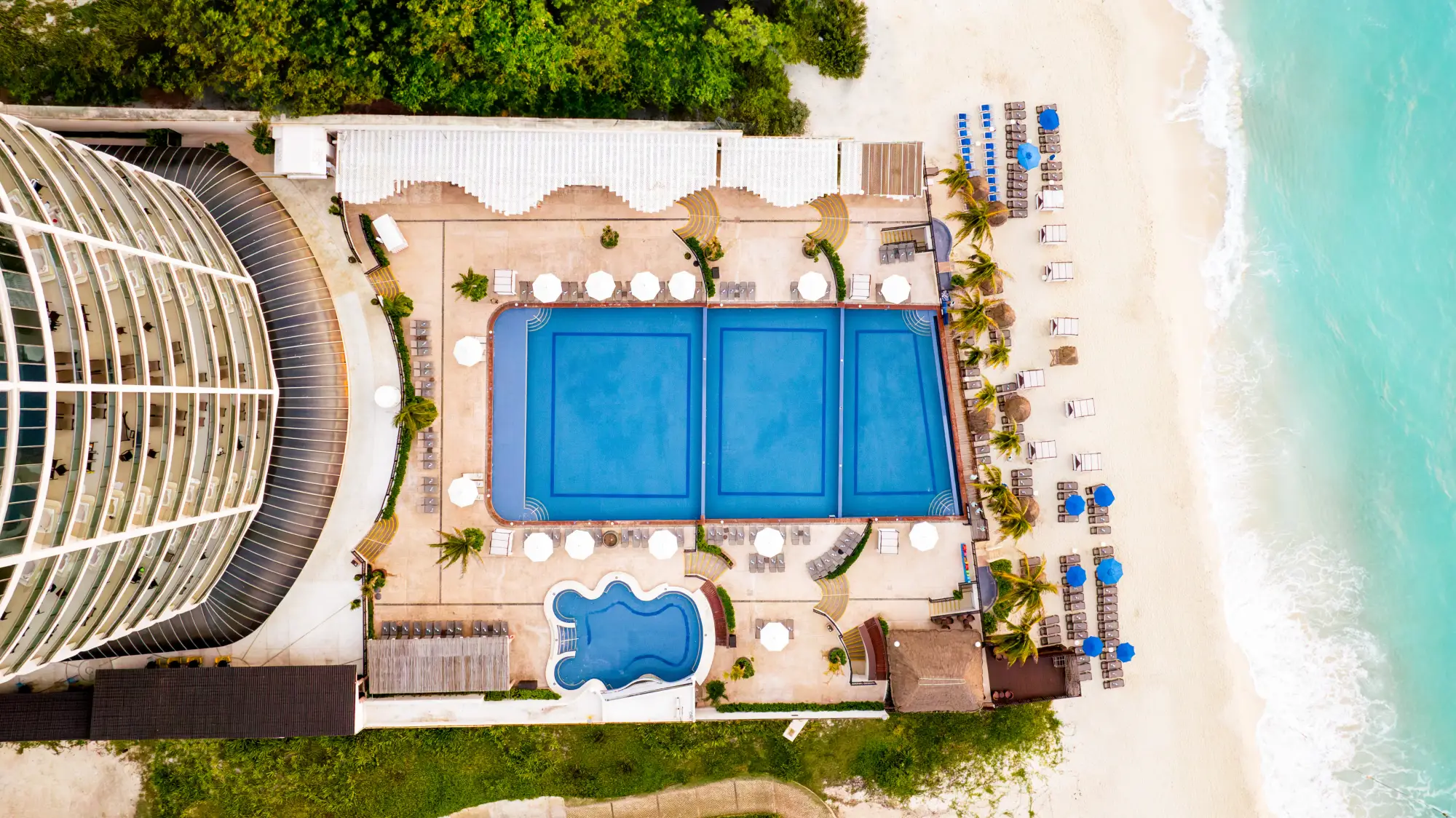 Aerial view of a beachside resort pool area, with loungers, palm trees, and ocean waves.