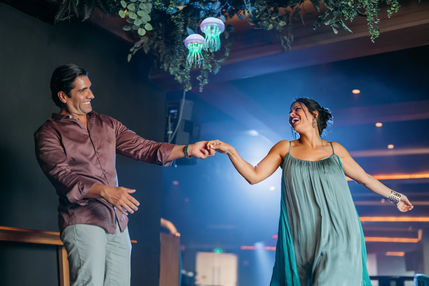 A smiling couple dances joyfully under hanging decor, in a softly lit room.