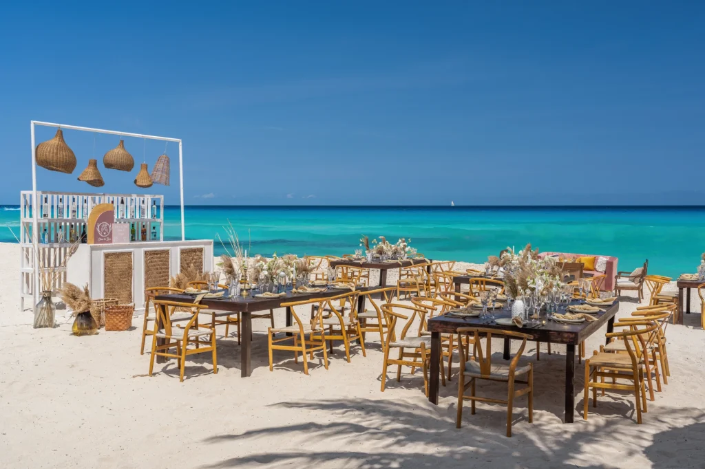 Beach wedding setup with wooden tables, chairs, and decorative lanterns on sand, overlooking turquoise ocean and clear sky.