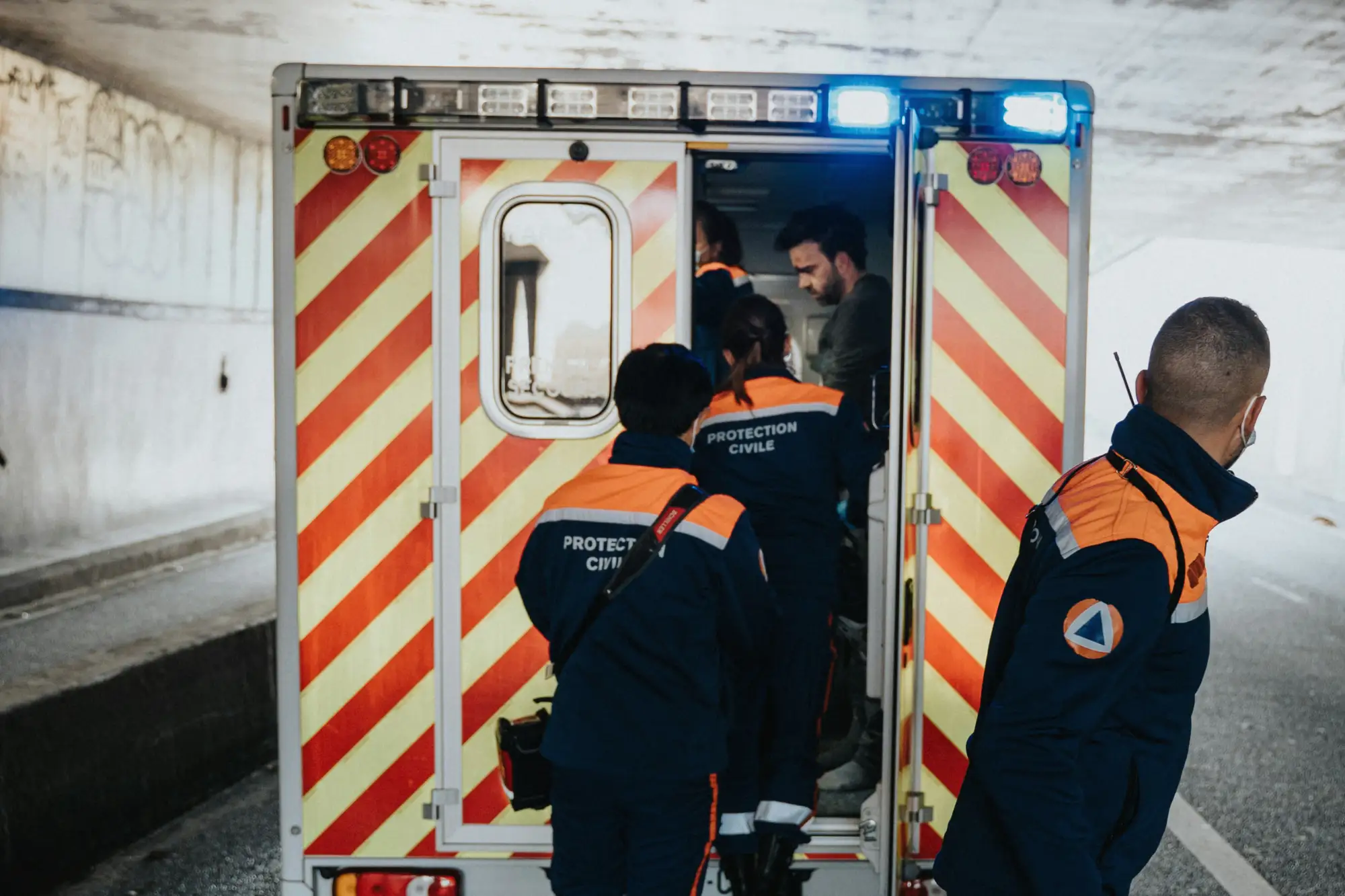 Rescue workers assisting a man into an ambulance under a bridge, with flashing lights on the vehicle.