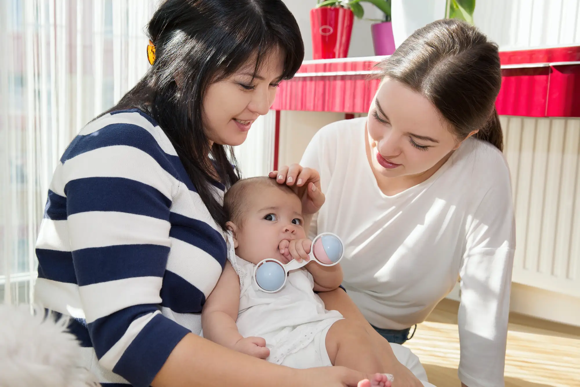 Two women smiling at a baby holding a rattle, sitting indoors with sunlight streaming in.
