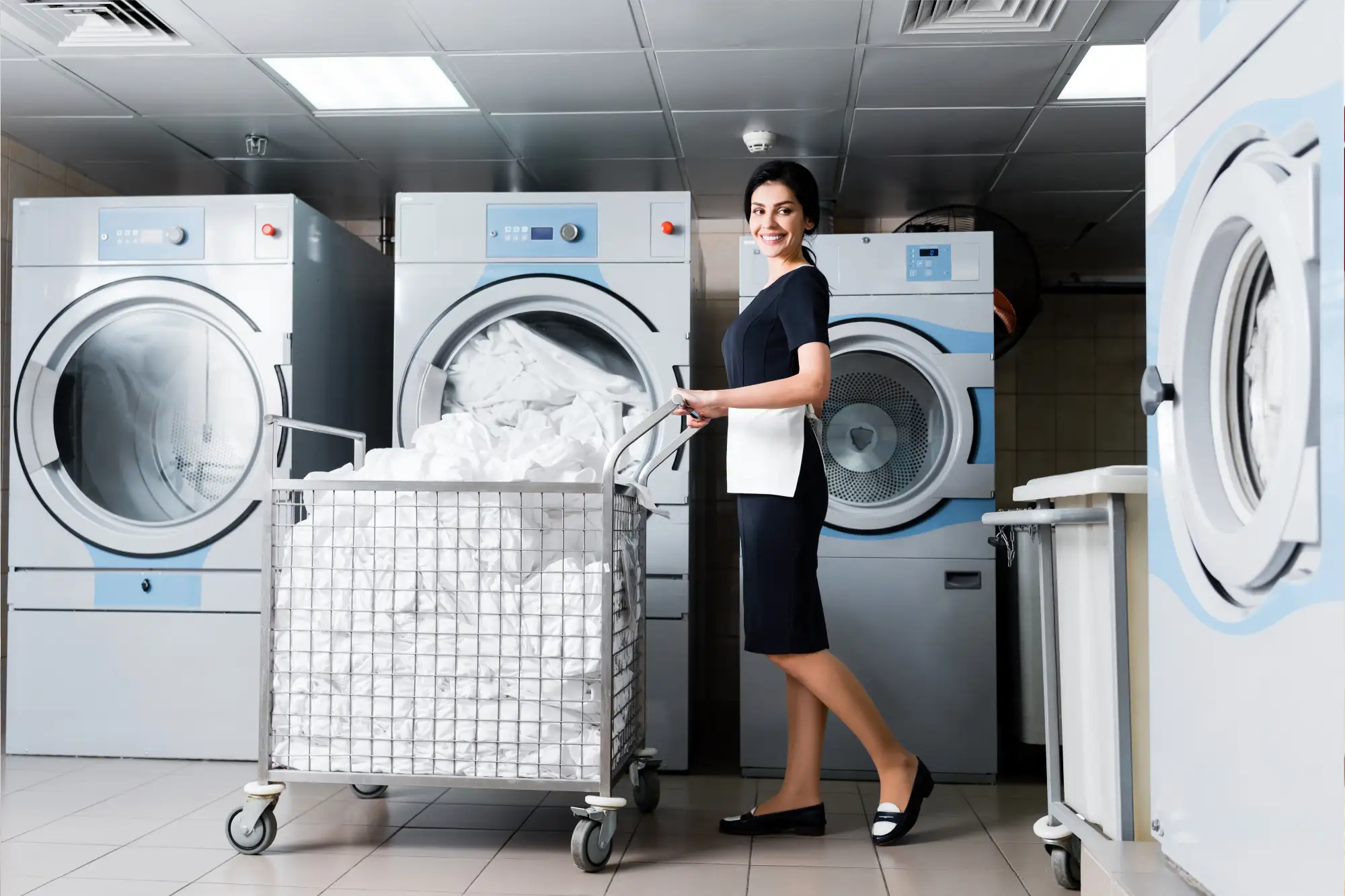 Woman in uniform smiling, pushing laundry cart filled with towels, standing by industrial washing machines in laundry room.