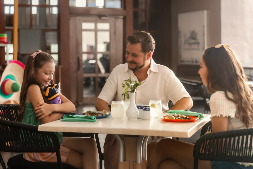 Family dining together at a restaurant, smiling and enjoying their meal around a table with drinks and colorful plates.