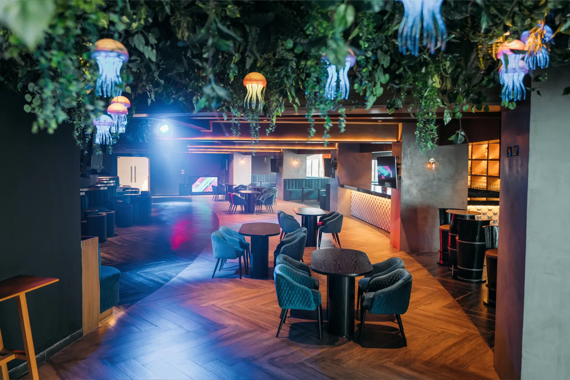 Stylish lounge with colorful jellyfish lights, elegant seating, and a modern bar, featuring abundant greenery.