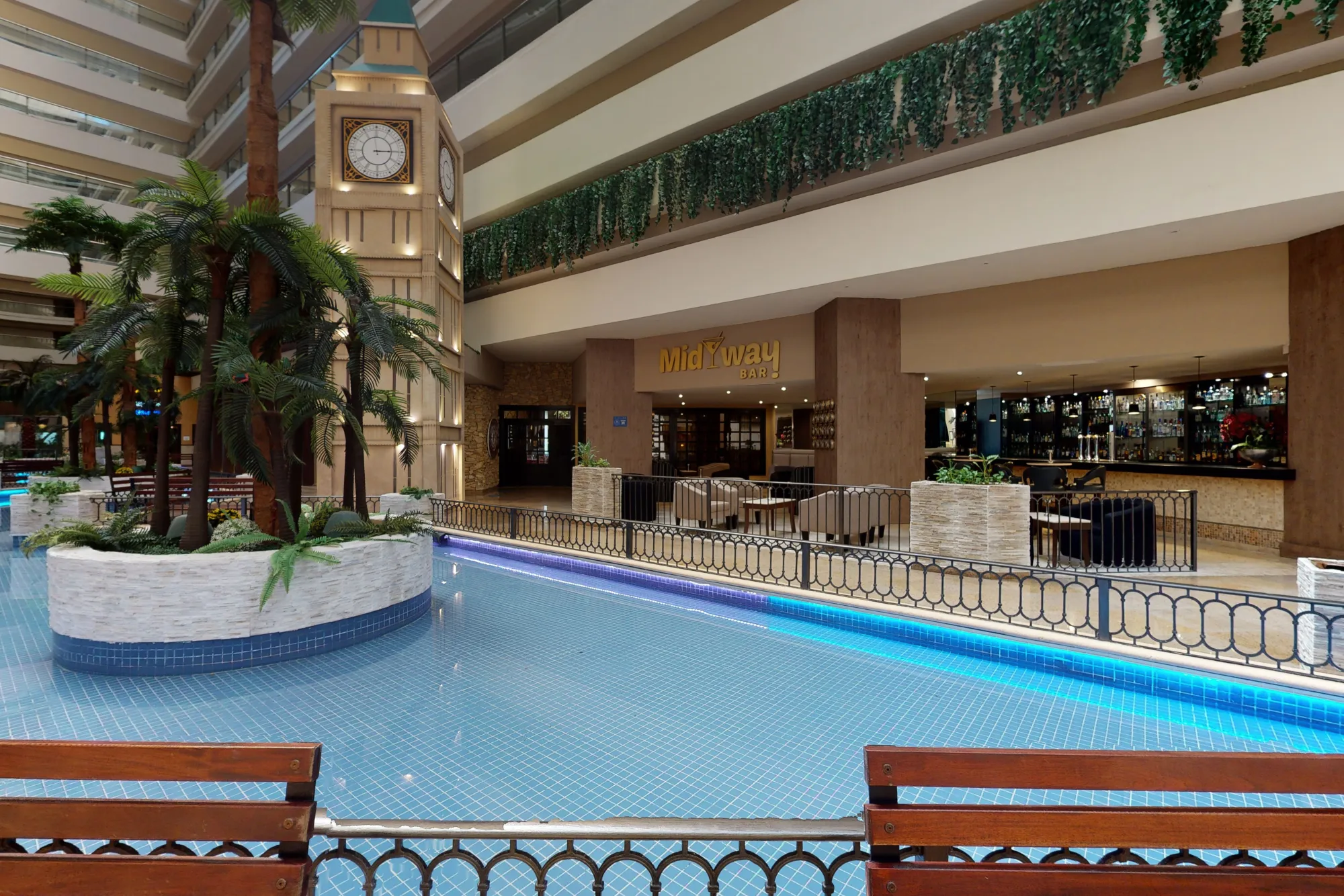 Indoor atrium with palm trees, a large clock tower, blue-tiled water feature, and the entrance to Midway Bar.