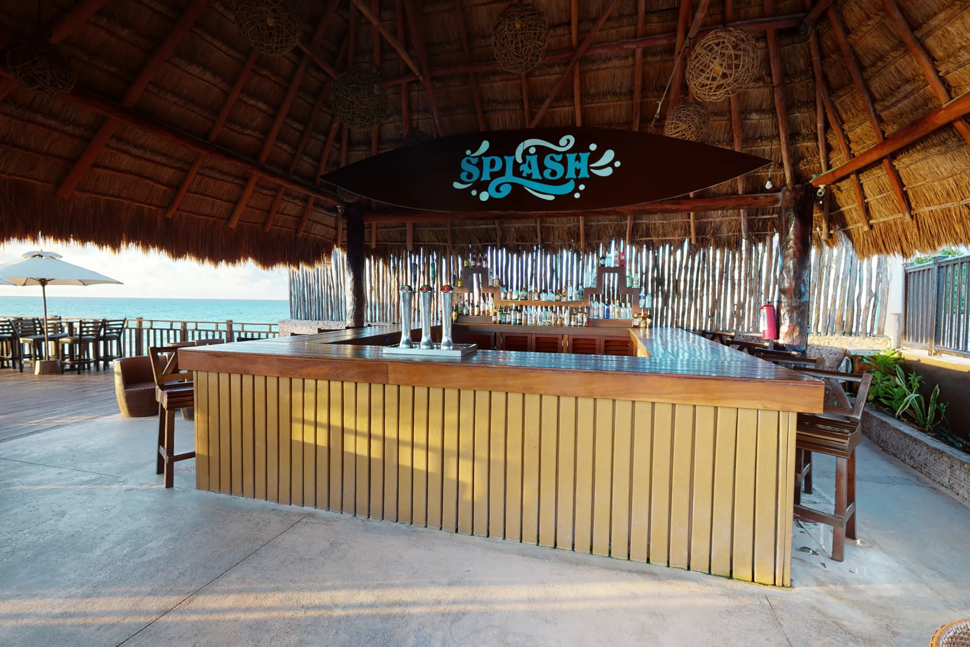 Tropical beach bar with a wooden counter under a thatched roof, overlooking the sea with outdoor seating.