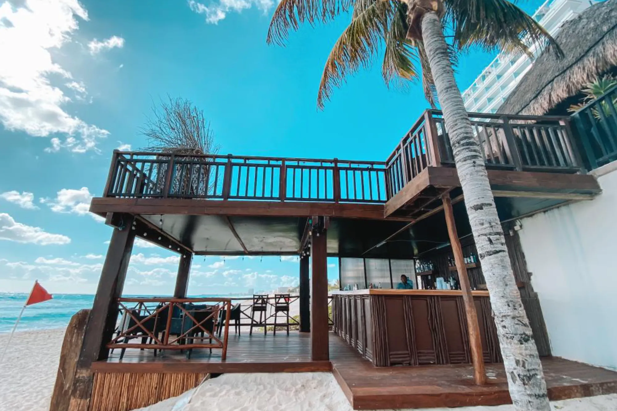 Beach bar with wooden deck and palm tree, set against a clear blue sky with ocean view.