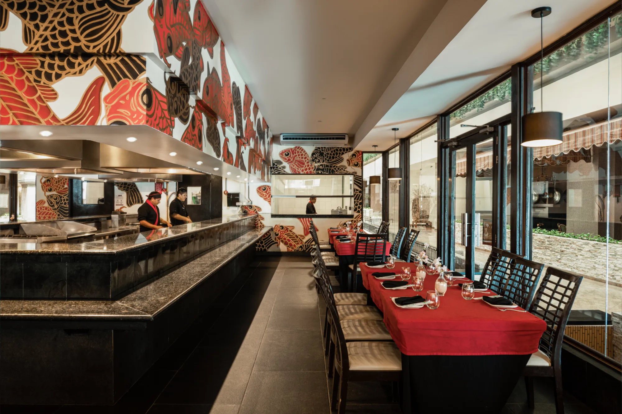 Restaurant interior with koi fish wall art, chefs behind a granite countertop, and tables set with red covers and black napkins.