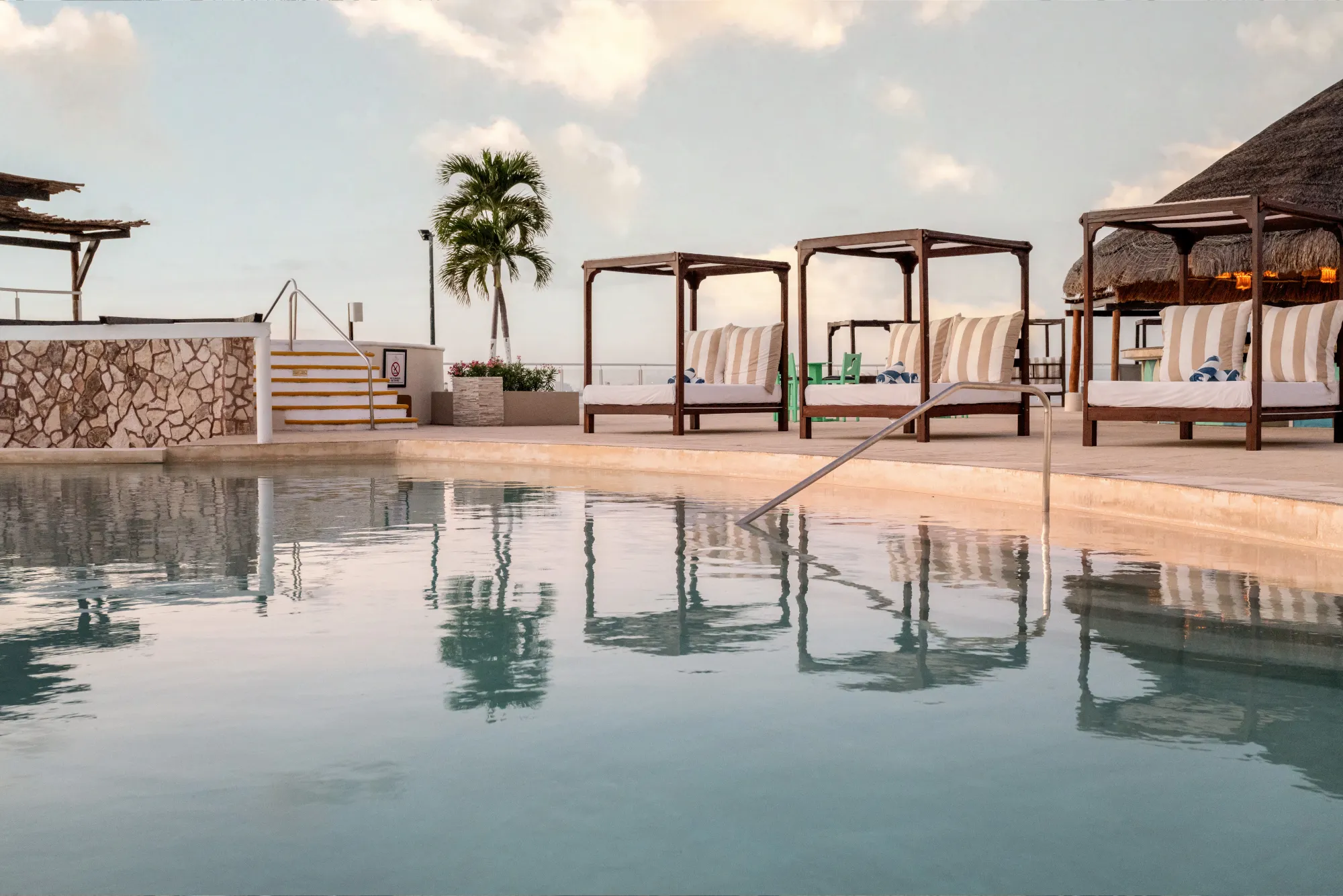 Poolside cabanas with striped cushions, a palm tree, and a thatched-roof area under a blue sky.