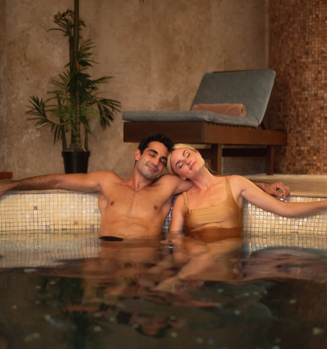 A couple relaxing in a cozy indoor pool, leaning against a tiled edge, surrounded by warm decor and a potted plant.