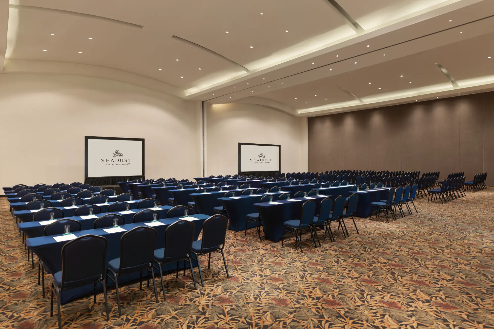 A conference room with rows of blue chairs and tables facing two large screens displaying "Seadust Cancun Family Resort."