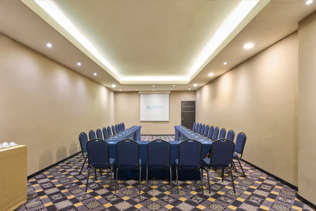 A U-shaped conference room with blue chairs, a beige carpet, and a screen at the front.