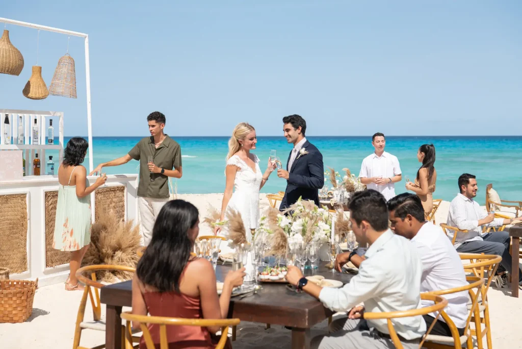 People enjoying a beach wedding reception with a decorated table and ocean view in the background.