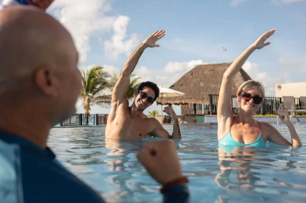 People doing arm exercises in a pool, with palm trees and a thatched roof in the background.