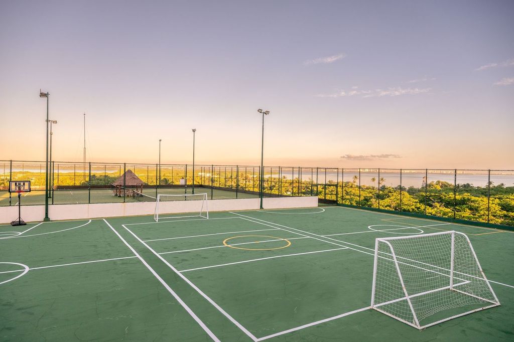 Outdoor sports court with goals, a basketball hoop, and lush greenery in the background under a clear sky.