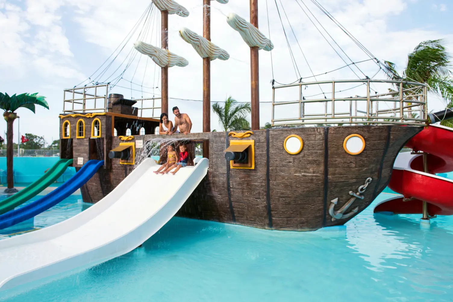 Children sliding down a pirate ship-themed water slide in a pool, with two adults watching from above.