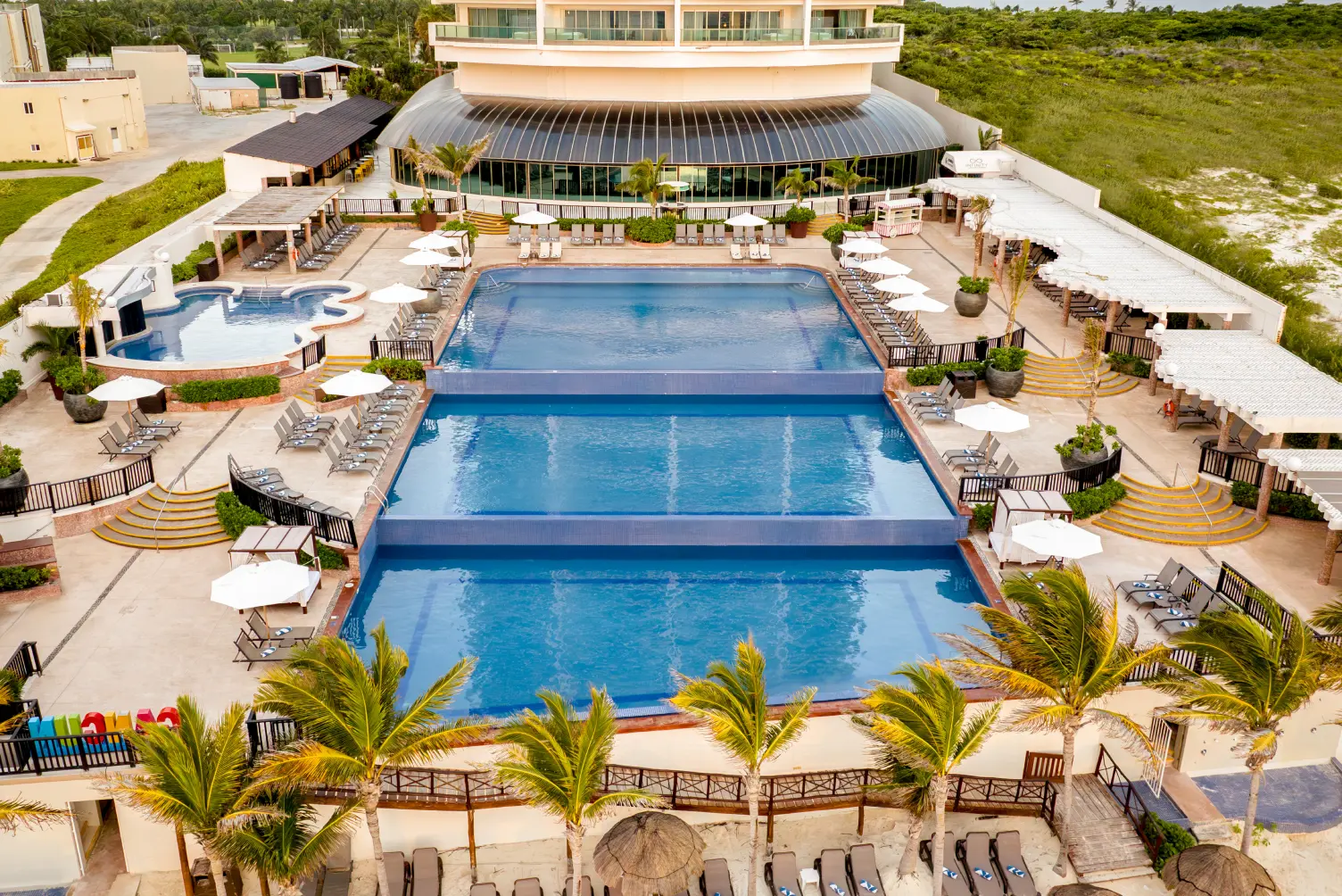 Aerial view of a hotel pool area with multiple pools, lounge chairs, umbrellas, and palm trees.