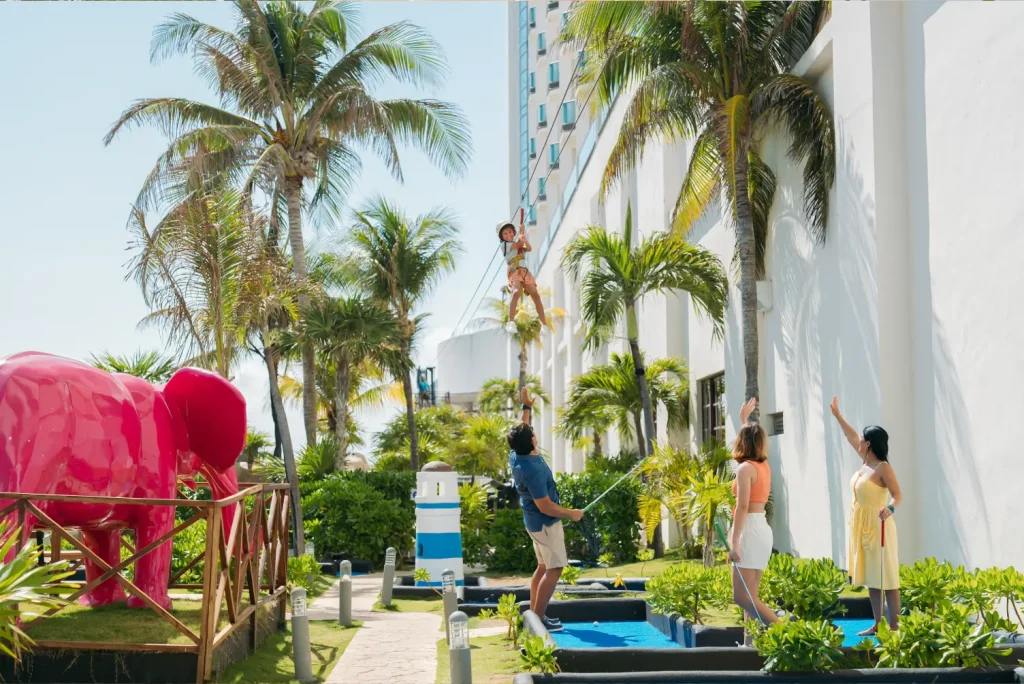 Child on zipline with adults guiding ropes, surrounded by palm trees and a large pink elephant statue nearby.