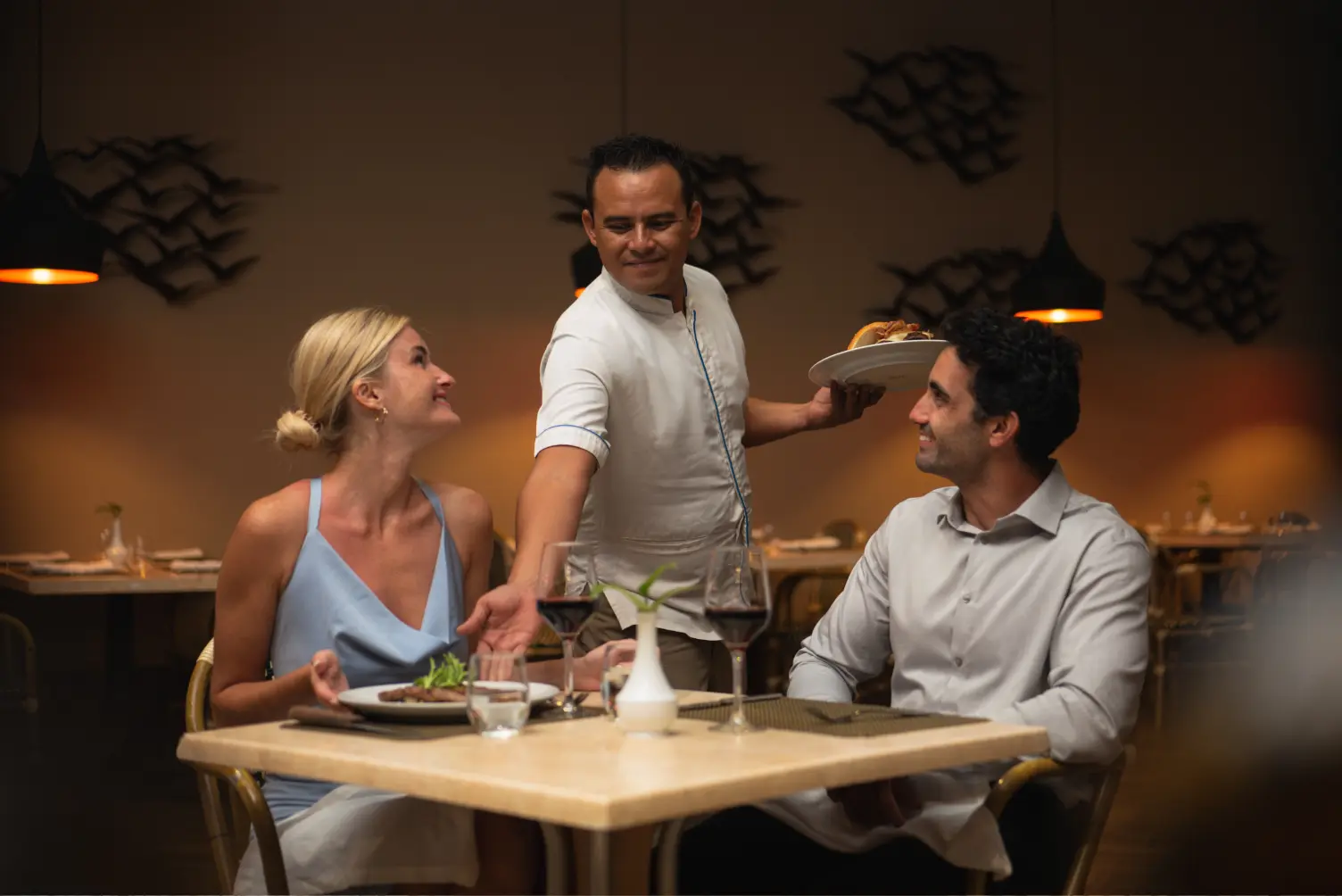 A couple in a restaurant is smiling as the waiter serves their meal under warm ambient lighting.