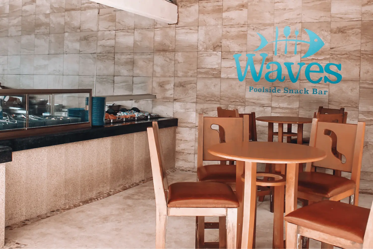Poolside snack bar with wooden tables and chairs, logo on tiled wall, counter with covered dishes on left.