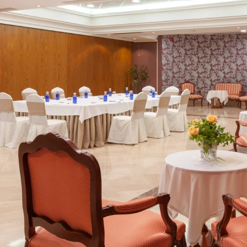 Sala de reuniones con sillas cubiertas, mesas decoradas con botellas azules y jarrón de flores sobre una mesa.