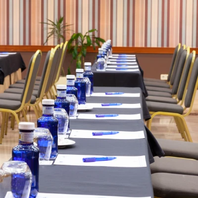 Mesas de conferencia con botellas de agua, blocs y bolígrafos en una sala con sillas alineadas.