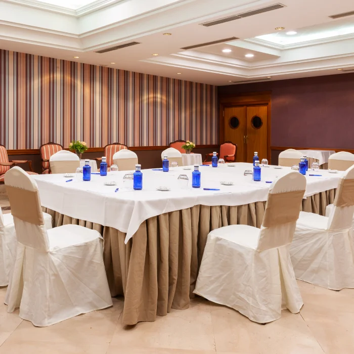 Sala de conferencias vacía con mesa cuadrada vestida, sillas cubiertas, botellas de agua y bolígrafos azules.