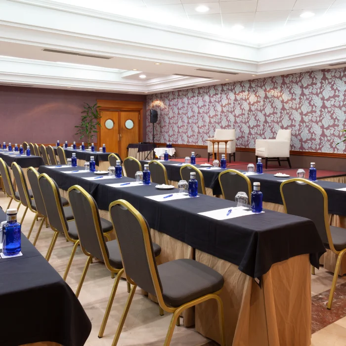 Sala de conferencias con sillas alineadas, mesas con manteles negros, libretas, bolígrafos y botellas de agua.