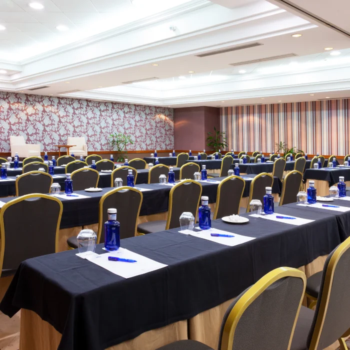 Sala de conferencias vacía con mesas largas, agua embotellada y bolígrafos sobre manteles oscuros. Decoración moderna.