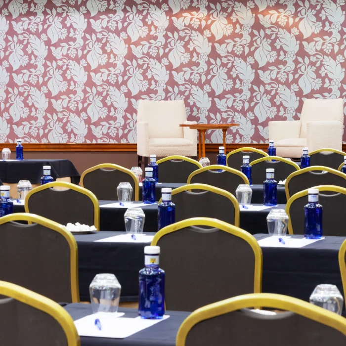 Sala de conferencias con sillas alineadas, botellas de agua y copas sobre las mesas, frente a un papel tapiz floral.