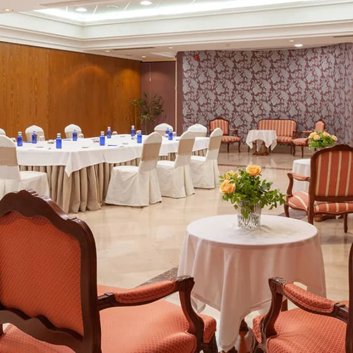 Salón elegante para eventos, con sillas y mesas cubiertas, botellas de agua, flores y decoración en tonos cálidos.