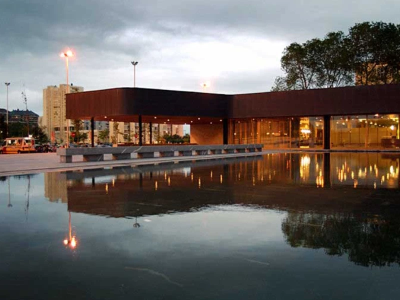Edificio moderno al atardecer reflejado en un estanque, con cielo nublado y árboles al fondo.
