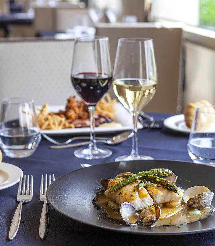 Plato gourmet con pescado, espárragos y almejas, acompañado de vino tinto y blanco en una mesa elegante.