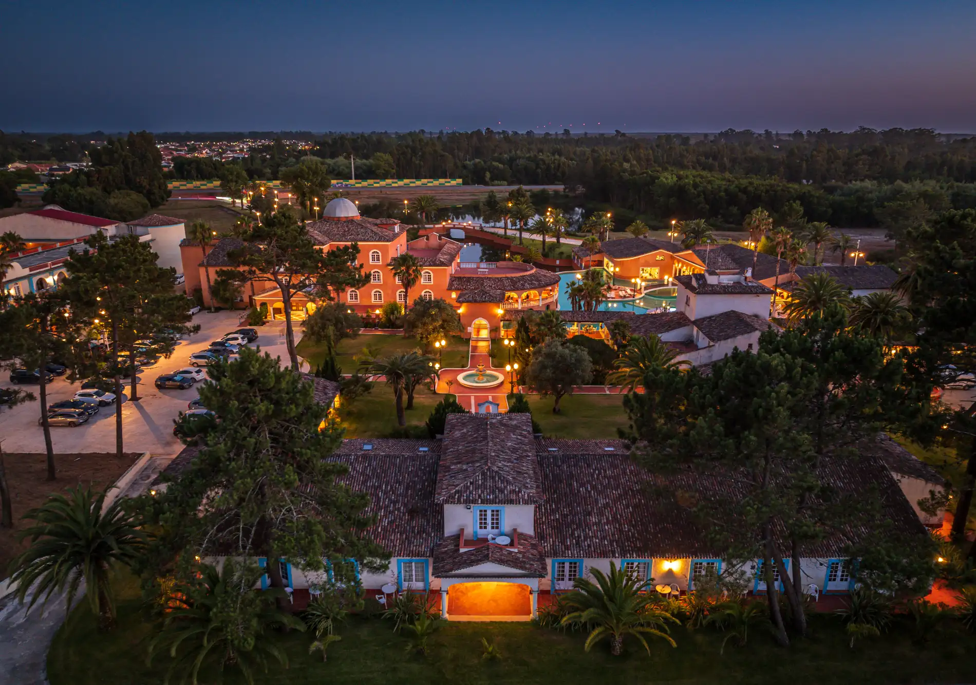 Aerial view of a lit-up hotel and pool at dusk, surrounded by trees.