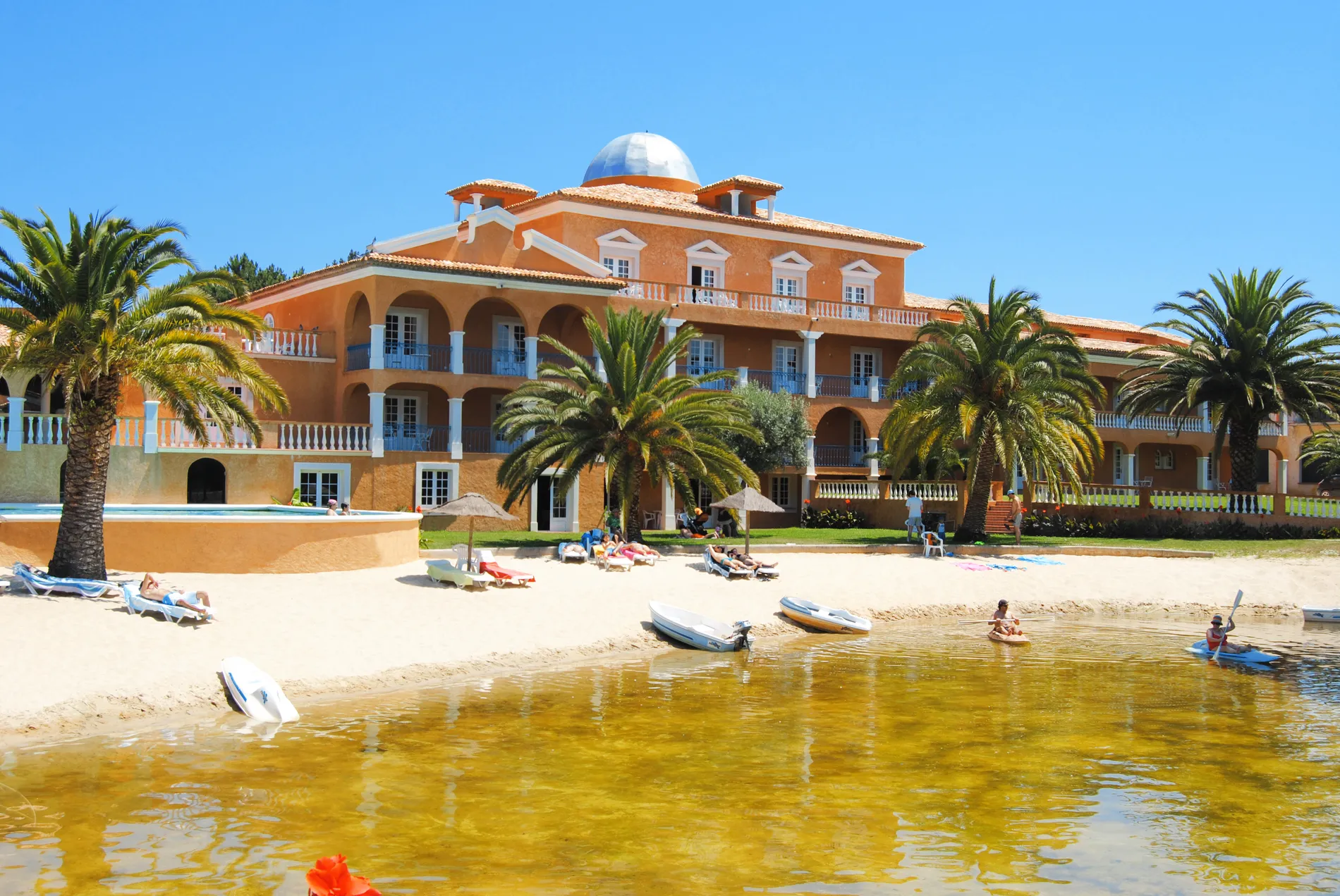 Beachfront hotel with palm trees, guests sunbathing and kayaking in the calm waters.
