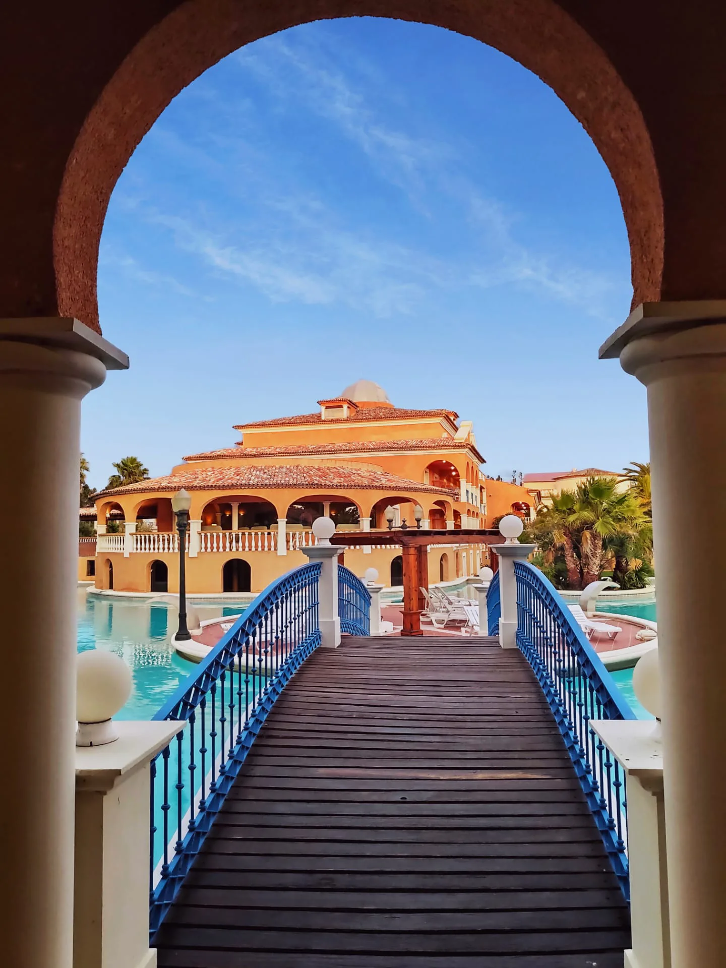 Arched walkway leading to a grand villa by the pool under a clear blue sky.