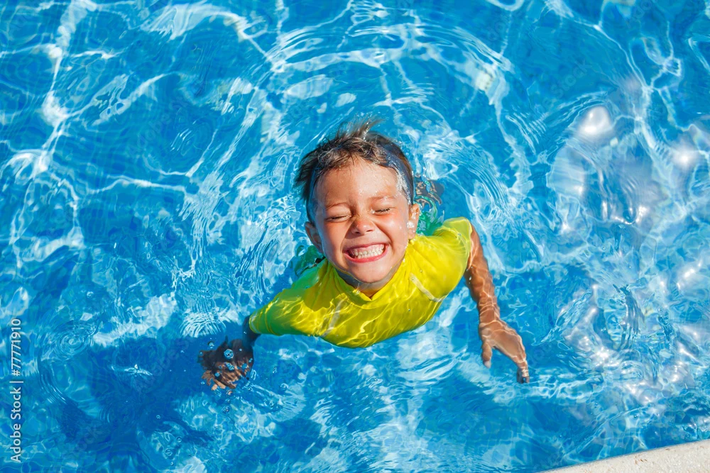 Child in yellow shirt grinning while swimming in a pool with clear blue water.