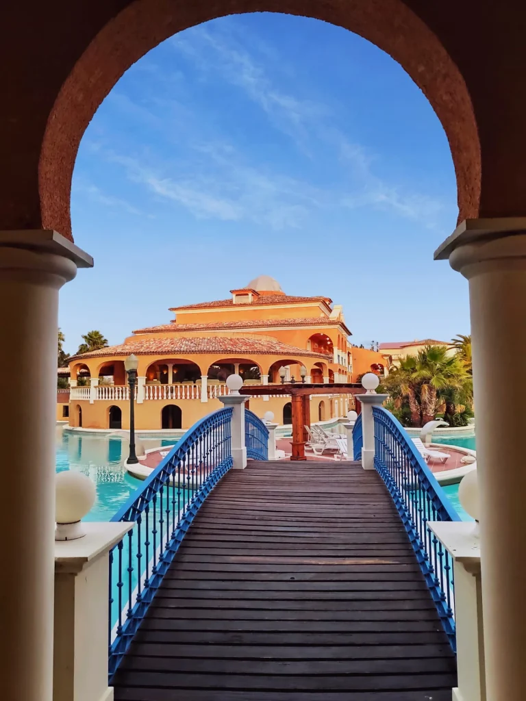 Arched view of a wooden bridge with blue railings leading to an ornate building by a pool under a clear sky.