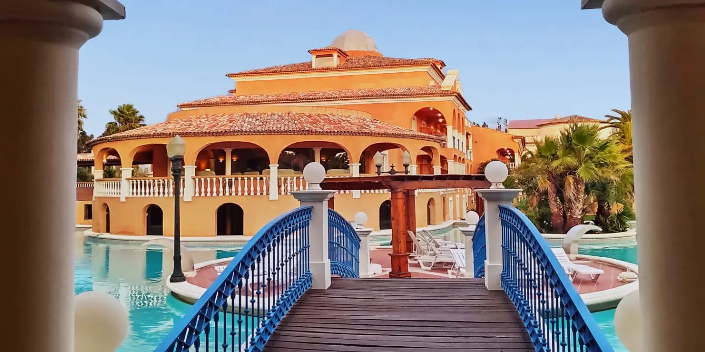 Bridge over a pool leading to a villa with palm trees and arches, under a clear blue sky.