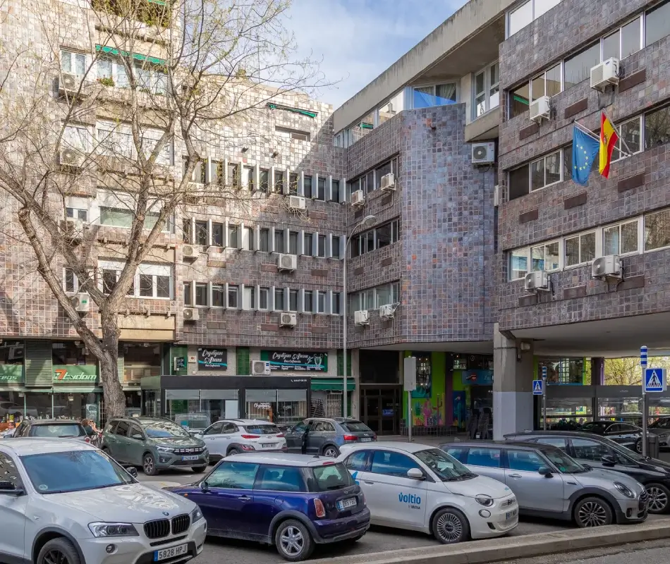 Edificio de oficinas con coches estacionados y árboles frente a él, bajo un cielo despejado.