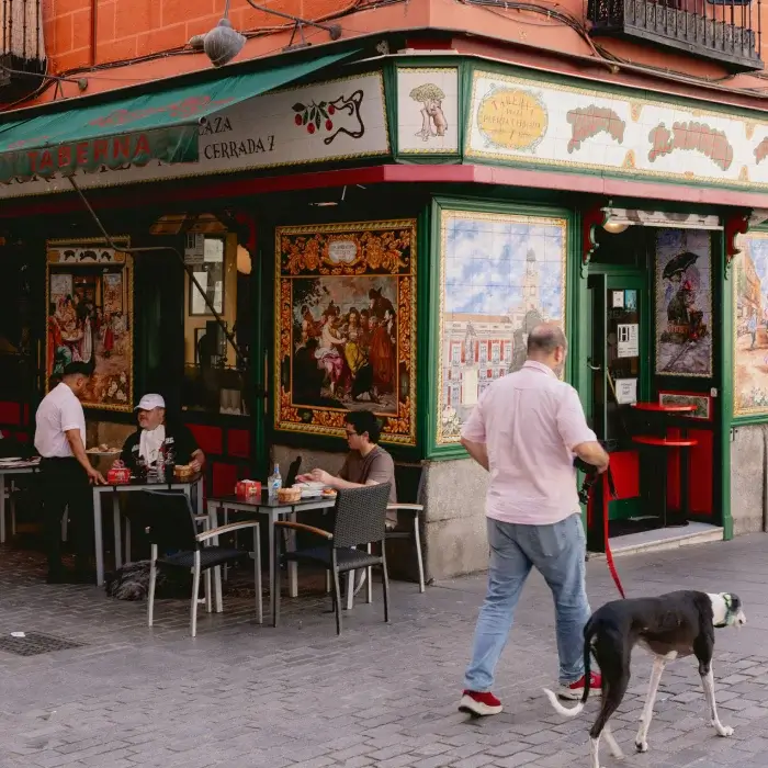 Hombre pasea a su perro cerca de una taberna con mesas al aire libre y azulejos decorativos.