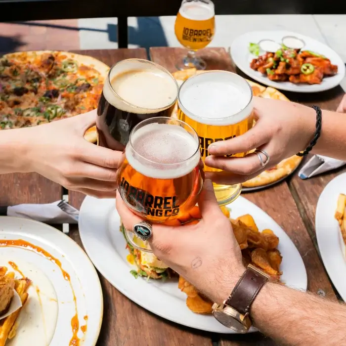 Cuatro manos brindando con cervezas sobre una mesa con pizza y alitas de pollo.