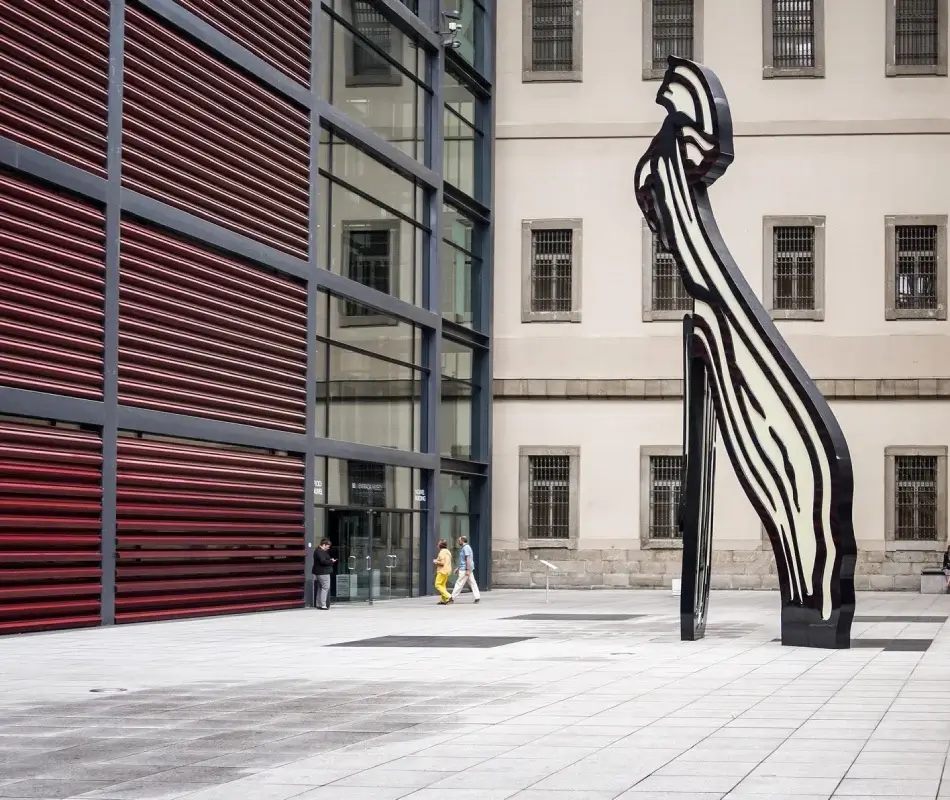 Escultura moderna junto a un edificio con paredes de vidrio y rejillas rojas. Personas caminando.