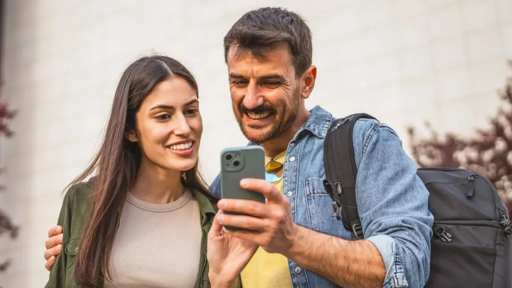 Pareja sonriendo mientras mira un teléfono móvil al aire libre.