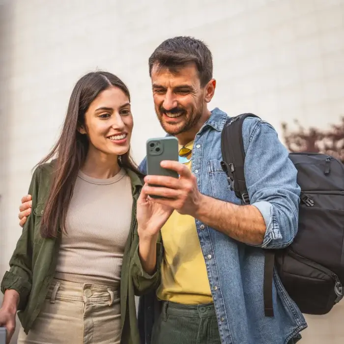 Pareja sonriendo mientras mira un teléfono móvil al aire libre.