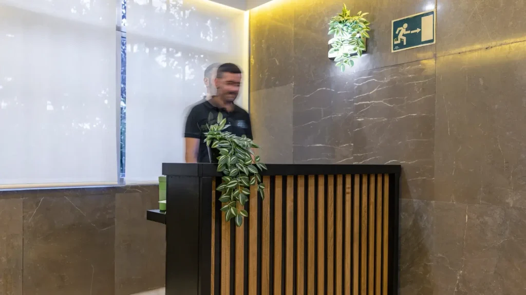 Persona borrosa detrás de un mostrador, decoración vegetal y señal de salida de emergencia.