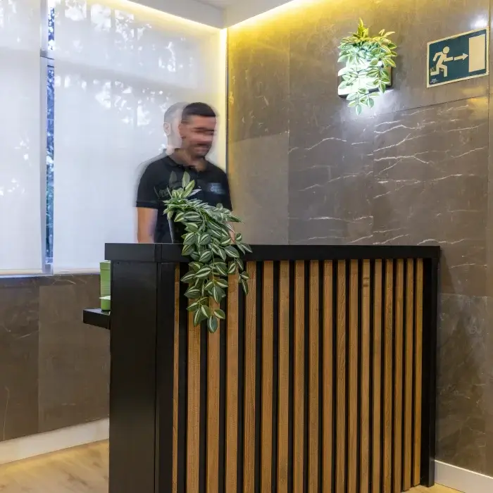 Persona detrás de un mostrador de recepción con plantas y señal de salida de emergencia.