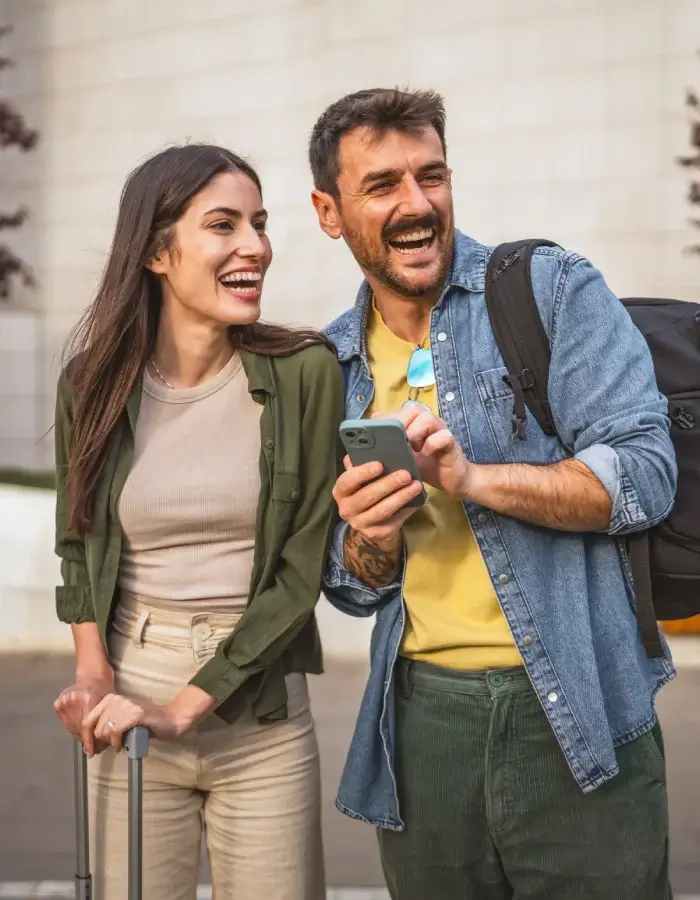 Pareja sonriente caminando con maleta y mochila mientras mira un teléfono móvil.