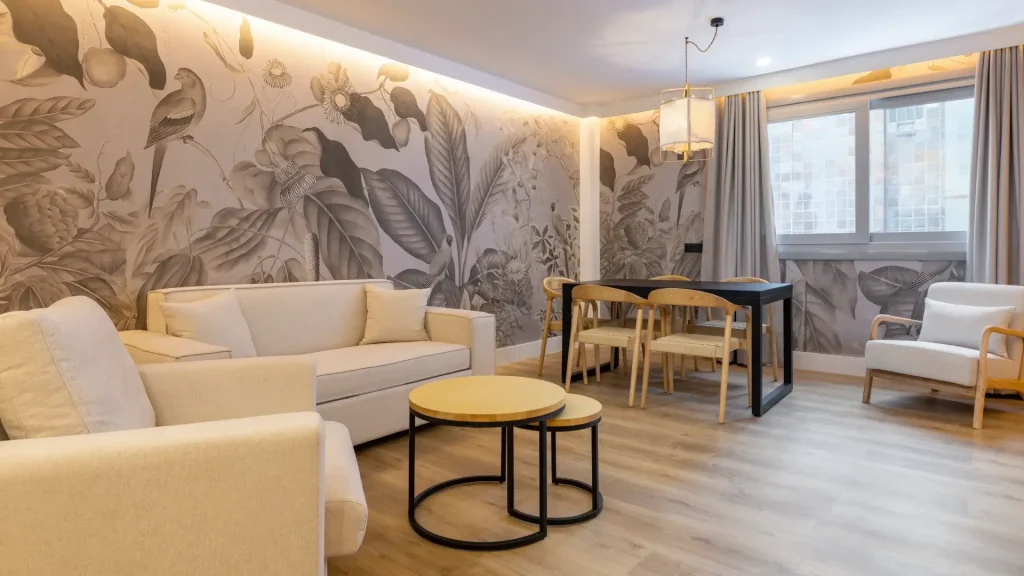 Sala moderna con muebles claros, mesas redondas y mural de hojas y aves en la pared.