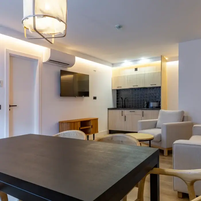 Sala moderna con kitchenette, mesa, sillones, televisor y lámpara colgante.